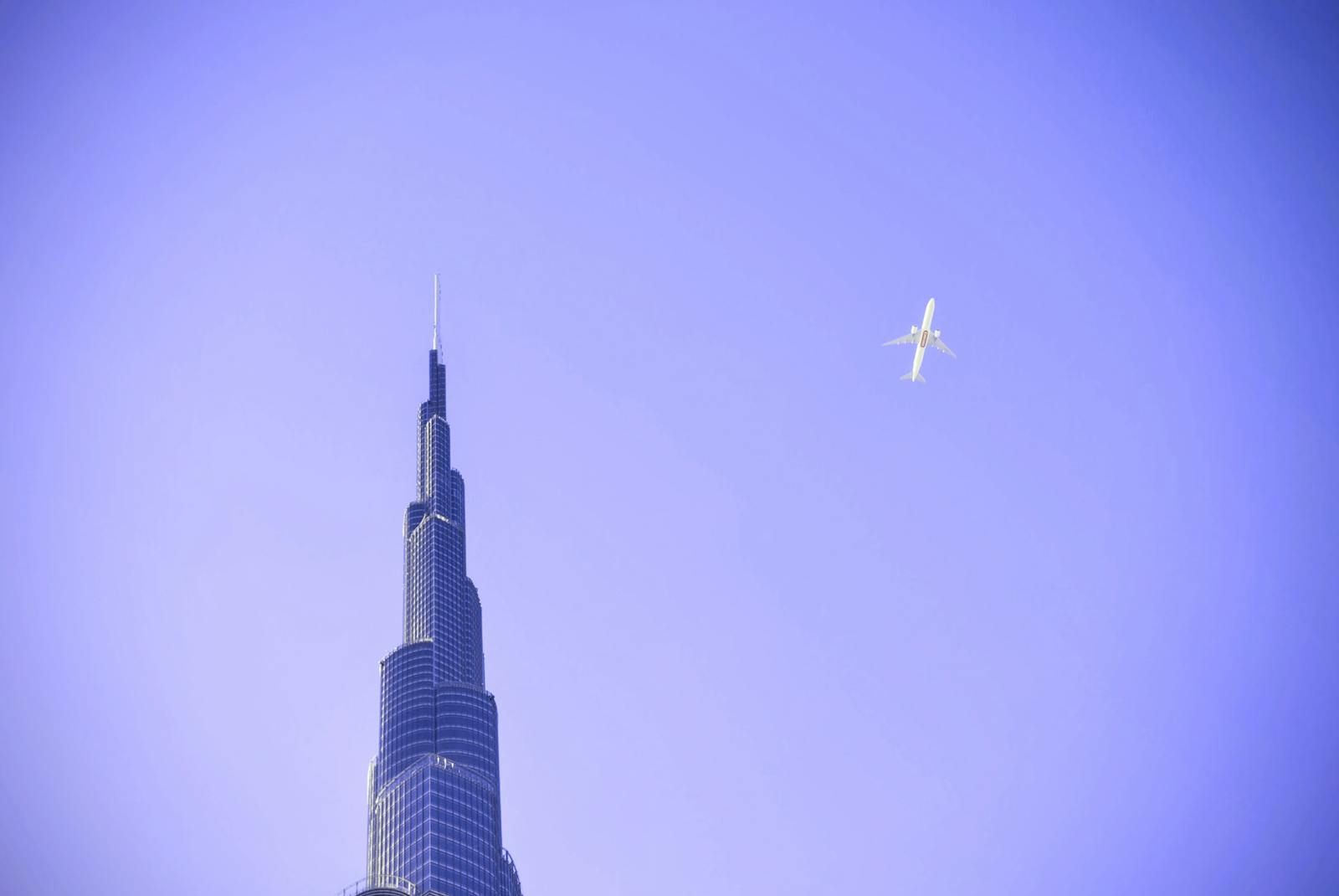 A modern airplane flies over the Burj Khalifa skyscraper against a clear blue sky in Dubai, UAE.